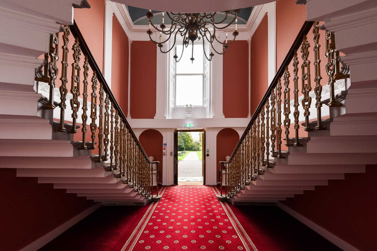 Grand staircase in Kilgraston House with ornate railings and a red carpet leading to an open doorway.