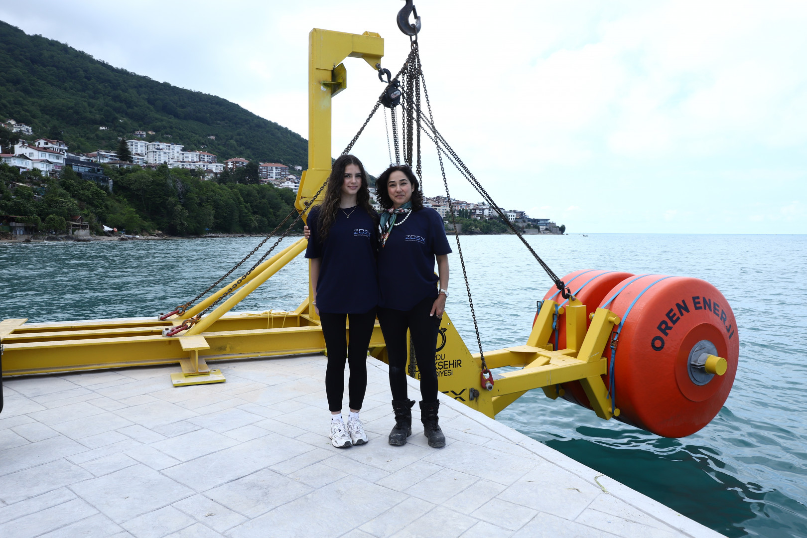 Two women stand near a yellow device being lowered into the water in Ordu, Turkey.